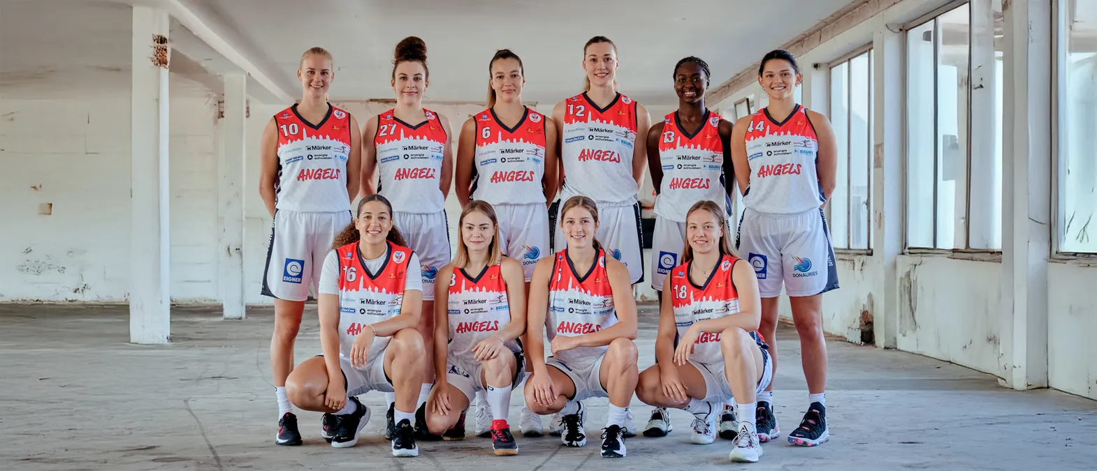 Een groep van acht vrouwen in oranje en witte basketbaltruien met de tekst 'ANGELS' poseren in een magazijn.