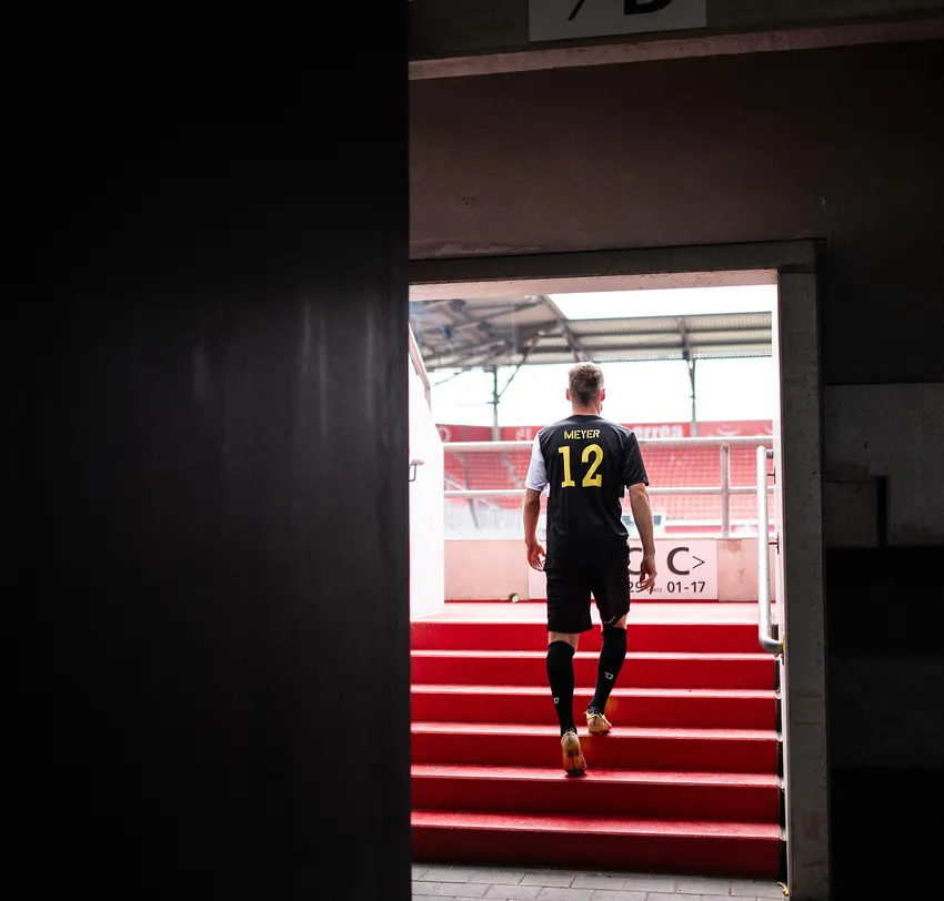 Een voetballer in een zwart shirt met 'MIEYER 12' in geel loopt de rode trappen van een stadion op.