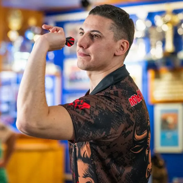 Een man in een gemusterd sportshirt met rode accenten houdt een dart vast en richt, met trofeeën op de achtergrond.