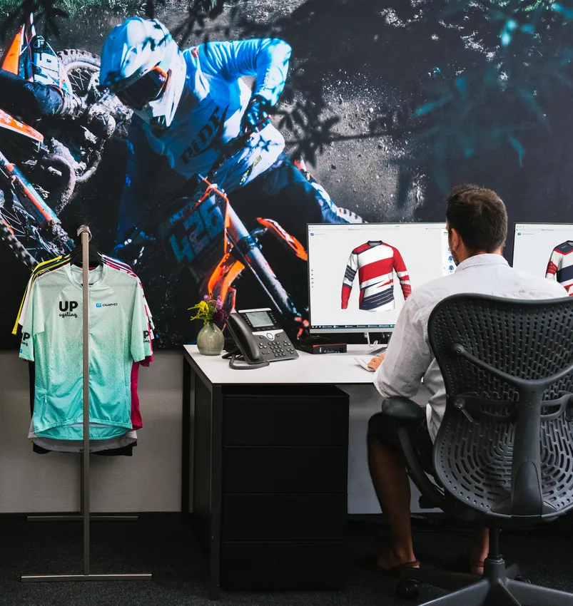 Een man zit aan een bureau en ontwerpt op een computer een aangepast sportshirt, terwijl op de achtergrond een groot beeld van een motorrijder en een kledingrek met diverse sportkleding te zien zijn.
