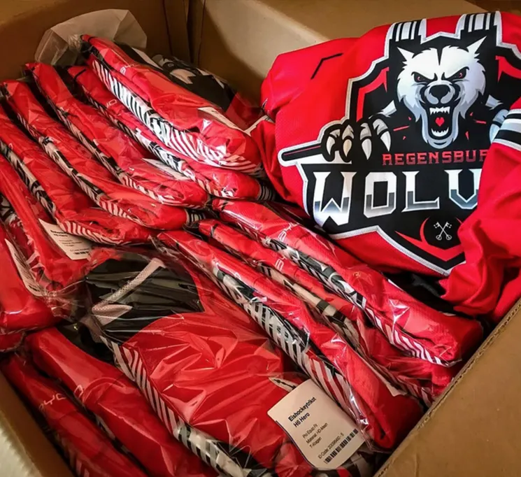 Een kartonnen doos gevuld met meerdere rode, op maat gemaakte sporttruien verpakt in plastic, waarvan er één een wolflogo en de tekst 'REGENSBURG WOLVES' heeft.