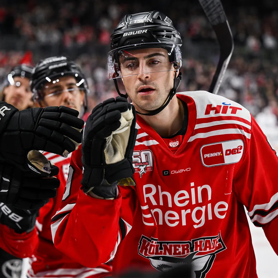 Een hockeyspeler in een rood shirt met witte strepen en logo's, die zijn hand opsteekt ter viering.