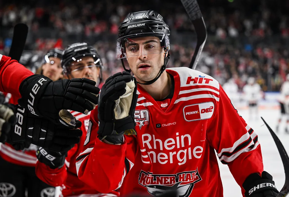 Een ijshockeyspeler in een rood shirt met zwarte handschoenen en een helm, die met teamgenoten viert.