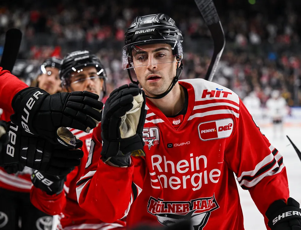 Een hockeyspeler draagt een rood shirt met witte en zwarte details terwijl hij met een teamgenoot interactie heeft.