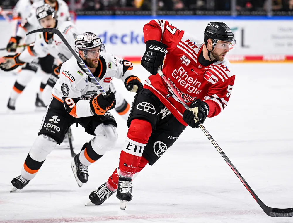 Een ijshockeyspeler in een rood shirt en zwarte broek schaatst over het ijs terwijl hij door een andere speler wordt achtervolgd.