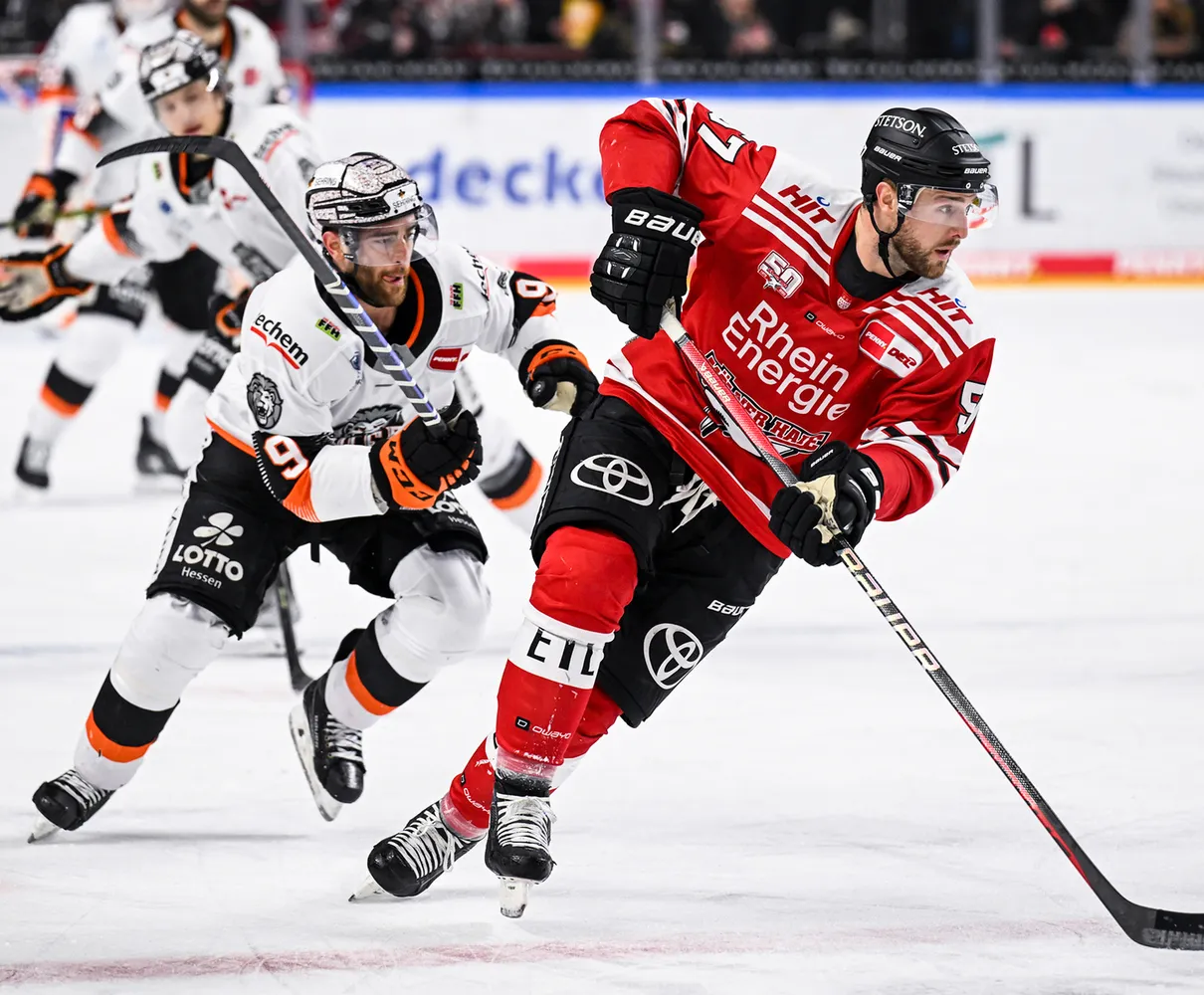 Twee ijshockeyspelers op het ijs, de ene in een rood shirt en de andere in een wit shirt met oranje accenten.