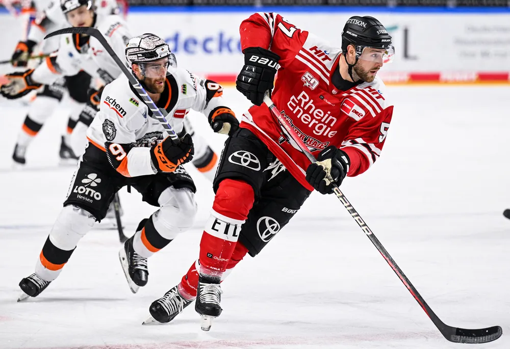 Een hockeyspeler in een rood shirt schaatst met de puck op het ijs terwijl een andere speler in een wit shirt dicht achter hem aan komt.