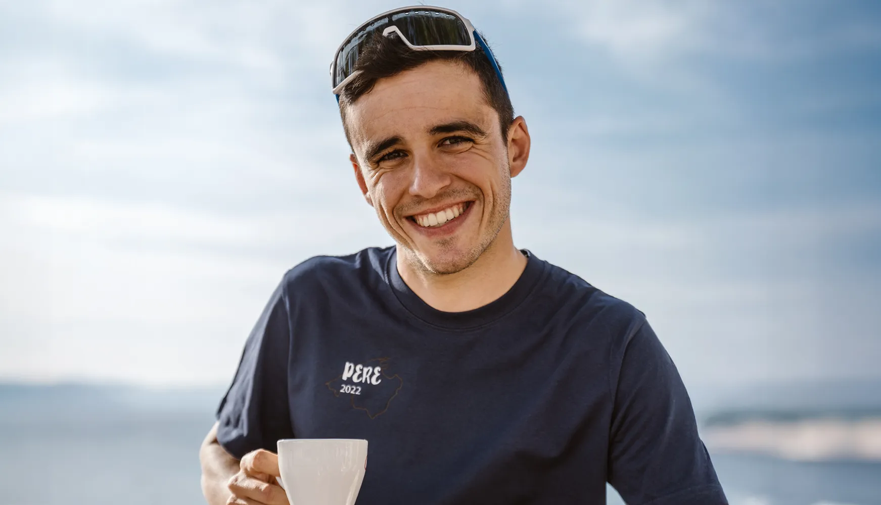 Een jonge man lacht in een blauwe T-shirt met de tekst 'PERE 2022' terwijl hij een kopje vasthoudt, tegen een heldere lucht.