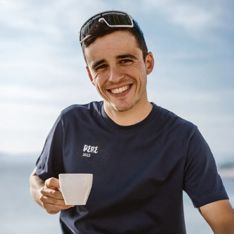 Een man houdt een kopje vast en draagt een donkerblauw T-shirt met de print 'PERE 2022', glimlachend voor een vervaagde achtergrond.