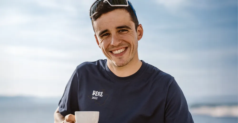 Een sportieve man in een marineblauw T-shirt met de opdruk 'PERE 2022' glimlacht terwijl hij een kopje vasthoudt, met een vage kustachtergrond.