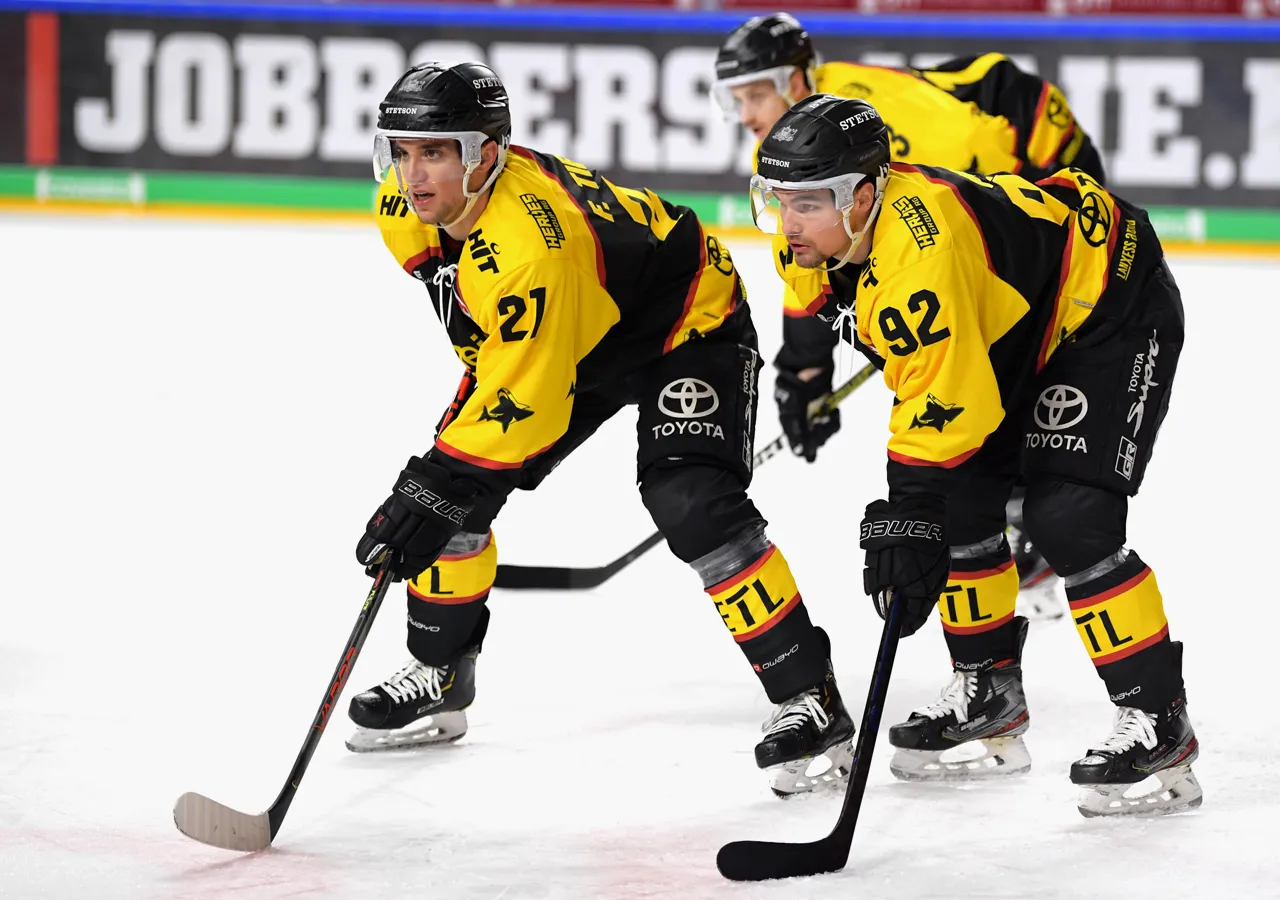 Twee ijshockeyspelers in gele en zwarte truien met nummers 21 en 92 op het ijs, geconcentreerd op het spel.