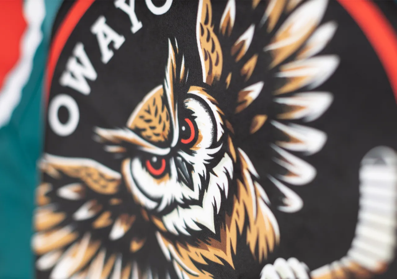 Een gestileerde uil met rode ogen op een zwarte achtergrond, omgeven door een rode en witte cirkel, op een sportkledingstuk van owayo.