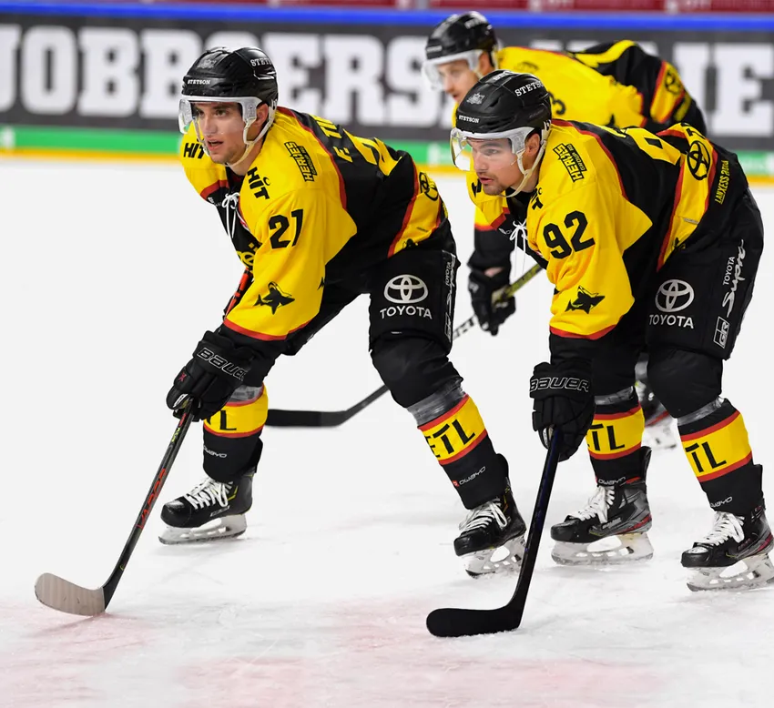 Hockeyspelers in gele en zwarte shirts met nummers 21 en 92 op het ijs, met hun stokken in de hand.