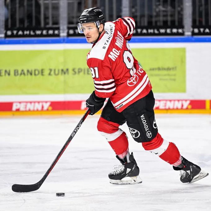 Een ijshockeyspeler in een rood en zwart shirt met nummer 91 en de naam Mc. Miller op de rug, staand op het ijs en een puck beheersend met zijn stok.