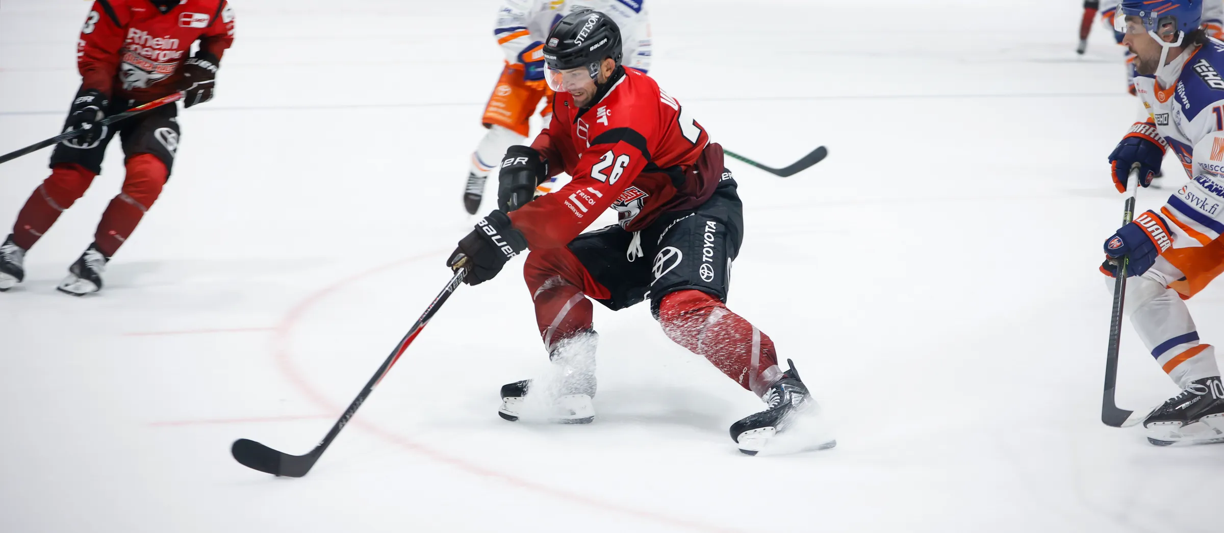 Een ijshockeyspeler in rode sportkleding dribbelt de puck op het ijs.