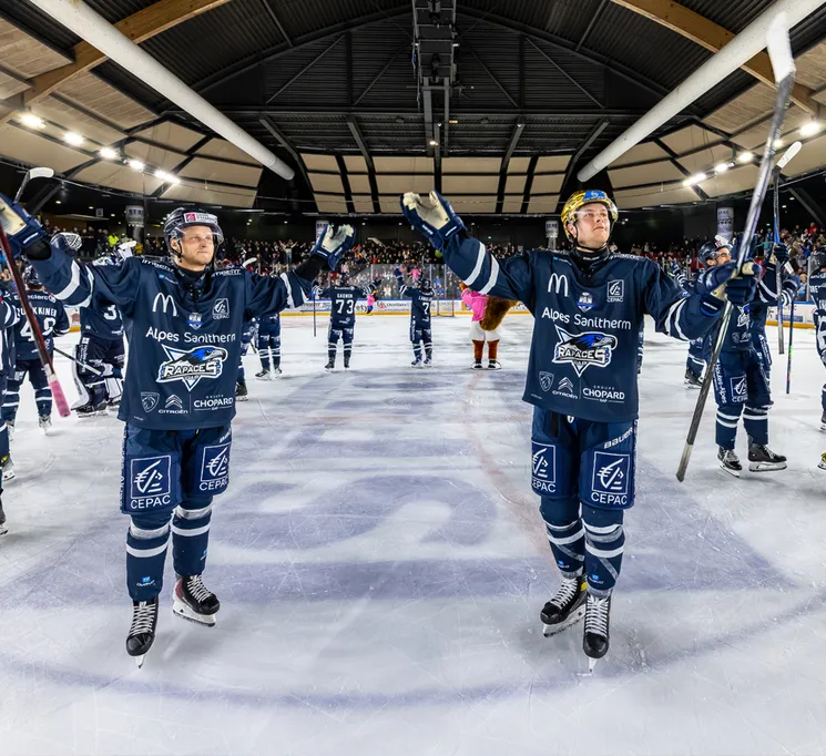 Hockeyspelers in blauwe shirts met sponsorlogo's vieren op het ijs, terwijl het publiek op de achtergrond zichtbaar is.