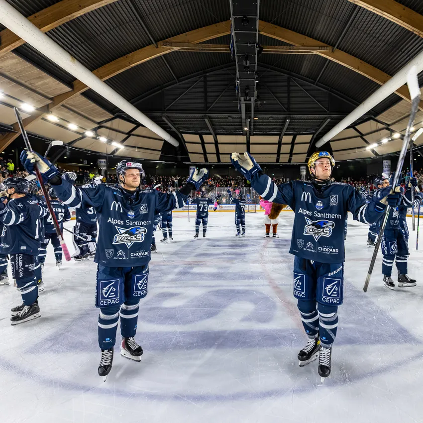 Twee ijshockeyspelers in blauwe shirts staan op het ijs en heffen hun stokken terwijl een juichende menigte op de achtergrond zichtbaar is.