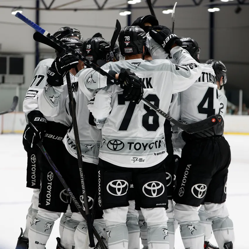 Een groep ijshockeyspelers in witte shirts omarmt elkaar op het ijs na het scoren van een doelpunt.