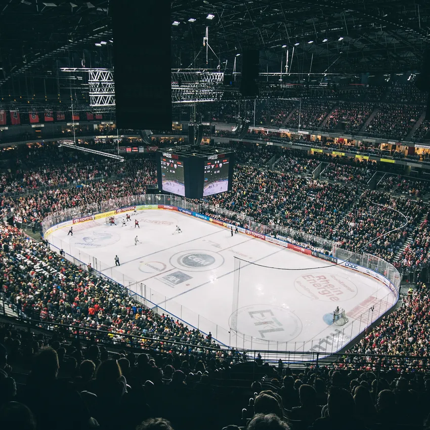 Een volle ijshockeyarena met juichende fans, een zichtbaar speelveld en een groot scherm op de achtergrond.