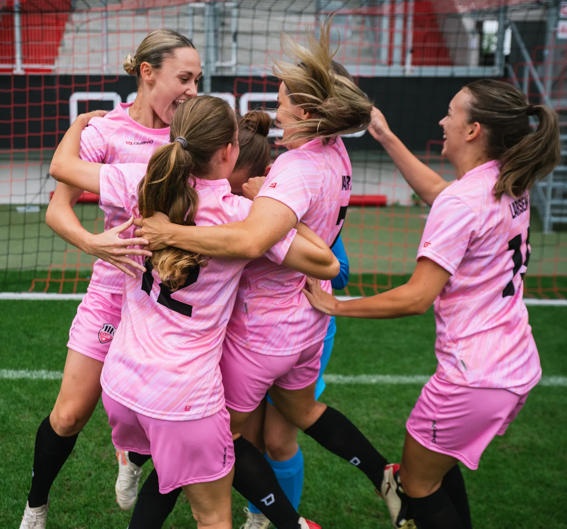 Vijf vrouwelijke spelers in roze shirts vieren samen vrolijk op het voetbalveld.