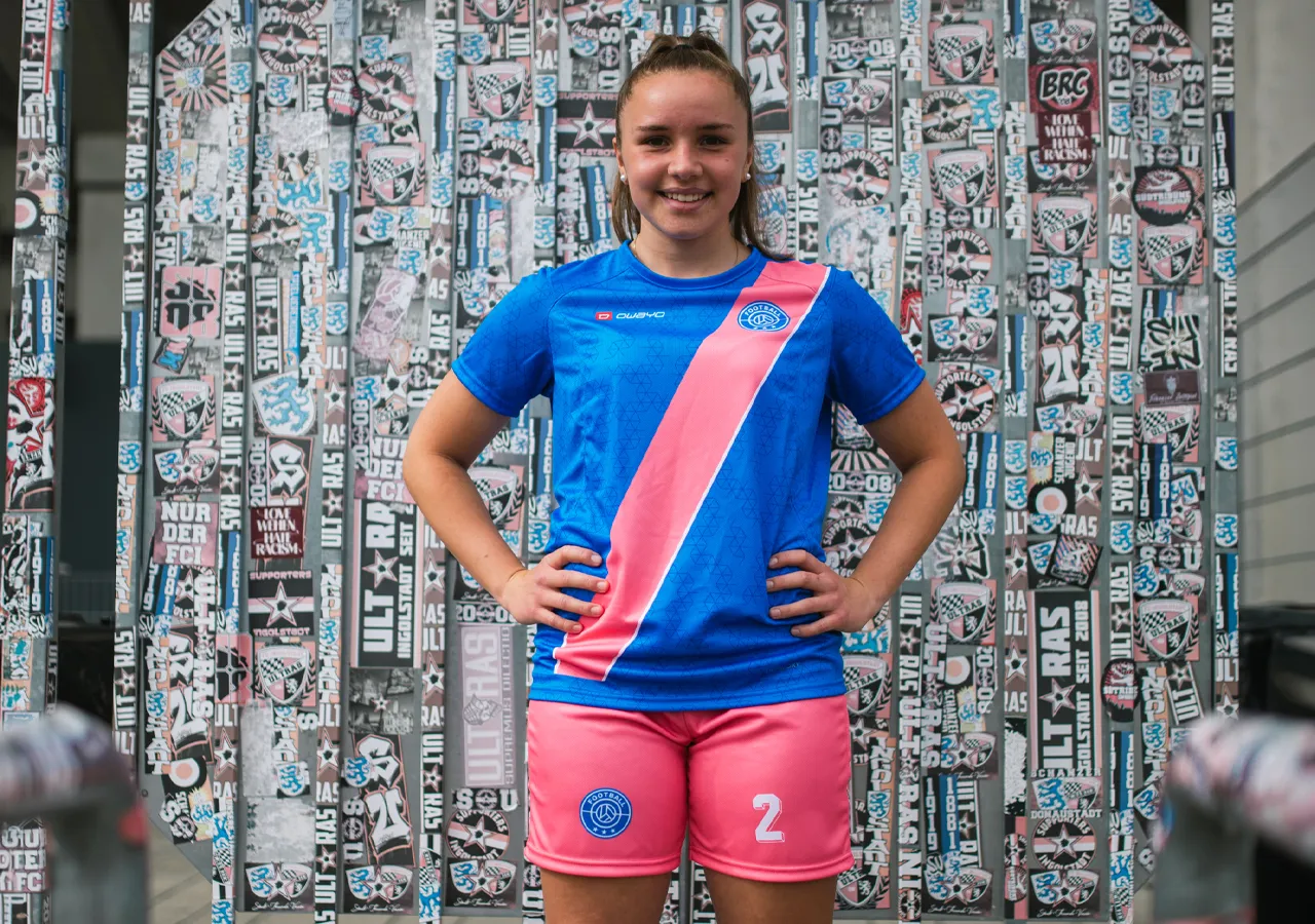 Een meisje in een blauw voetbalshirt met een roze diagonale streep en roze shorts, glimlachend voor een muur versierd met stickers.