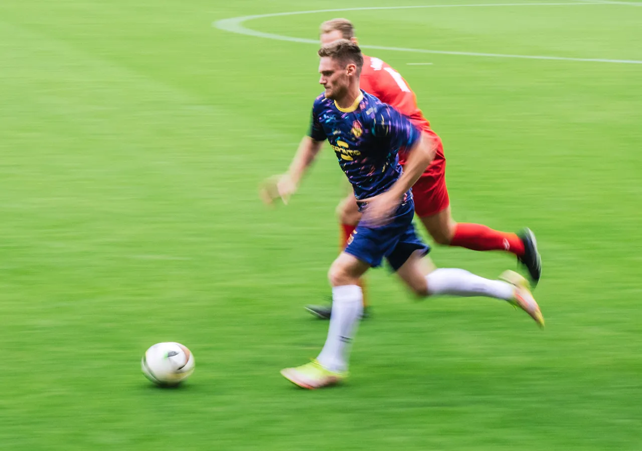 Een voetballer in een kleurrijk shirt sprint over het gras terwijl een andere speler dicht achter hem aanloopt.