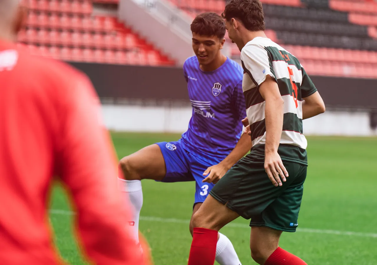 Twee voetballers in actie op het veld, de een draagt een blauw shirt en de ander een groen-wit gestreept shirt.