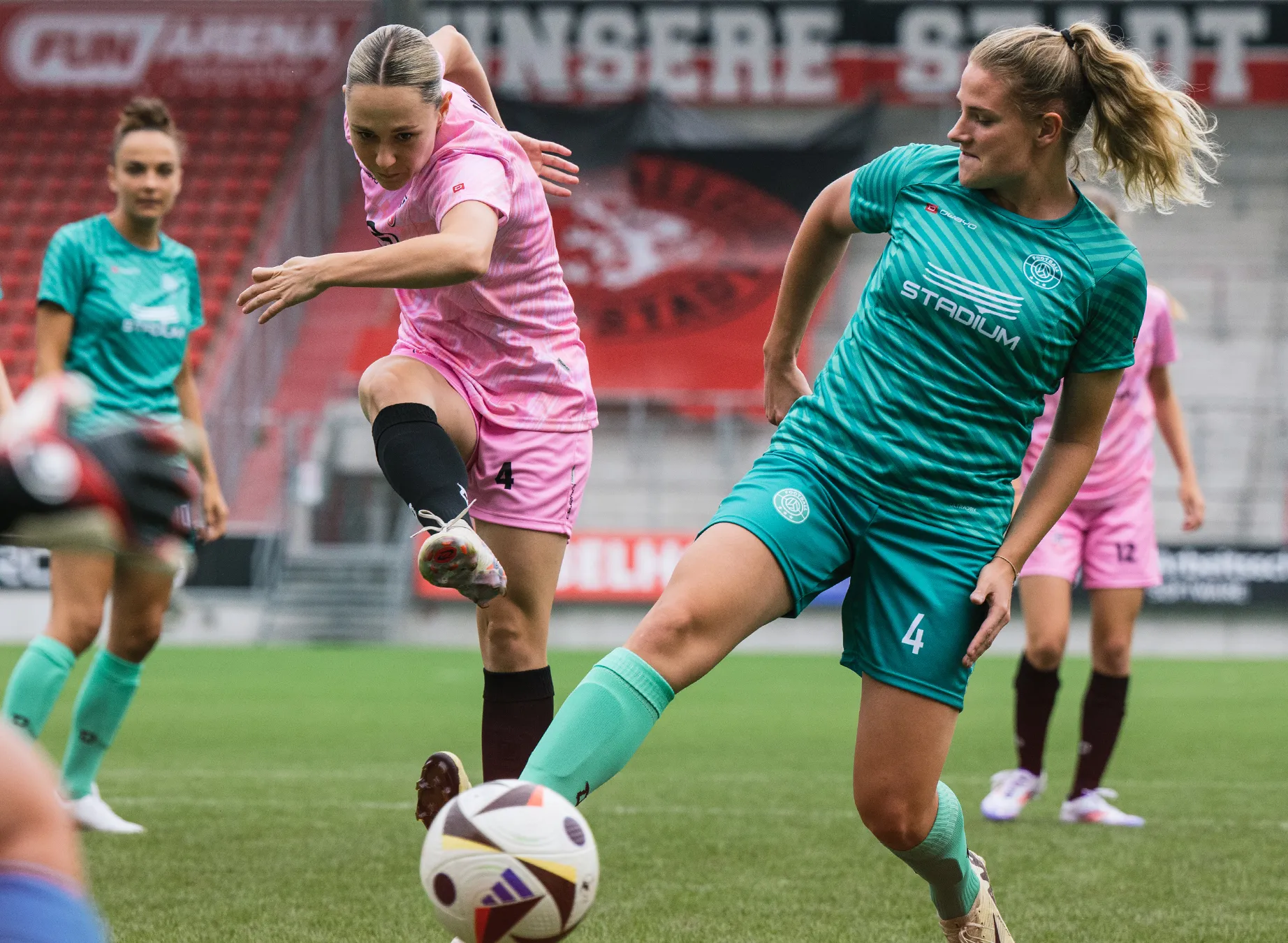 Twee vrouwelijke voetballers in levendige shirts, de een in roze en de ander in groen, spelen op een grasveld.