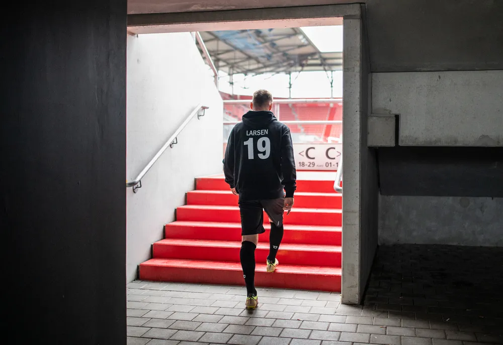 Een voetballer in een zwarte hoodie met het nummer 19 en shorts loopt de rode trappen van een stadion op.