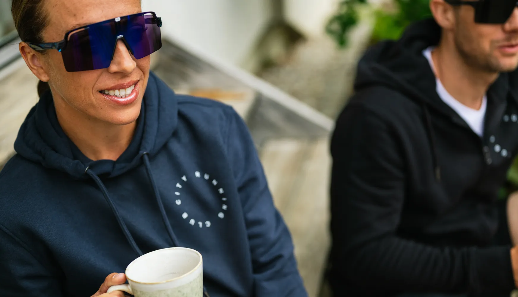 Een persoon in een blauwe hoodie houdt een beker vast en draagt grote zonnebrillen, terwijl een andere persoon op de achtergrond ook een hoodie draagt.