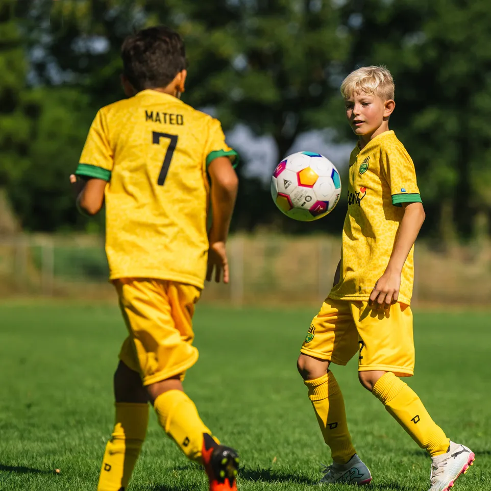 Twee kinderen in gele voetbalshirts en shorts spelen op een groen voetbalveld.