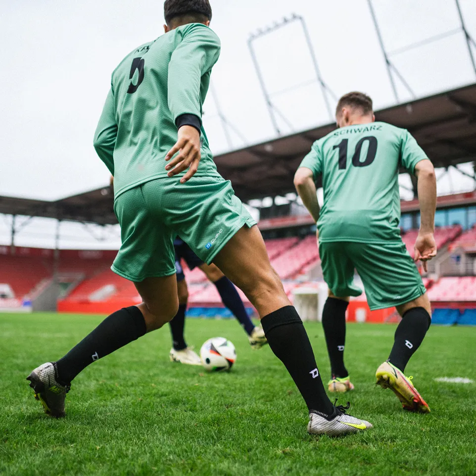 Twee voetballers in groene shirts en zwarte sokken die actief spelen op een grasveld in een stadion.