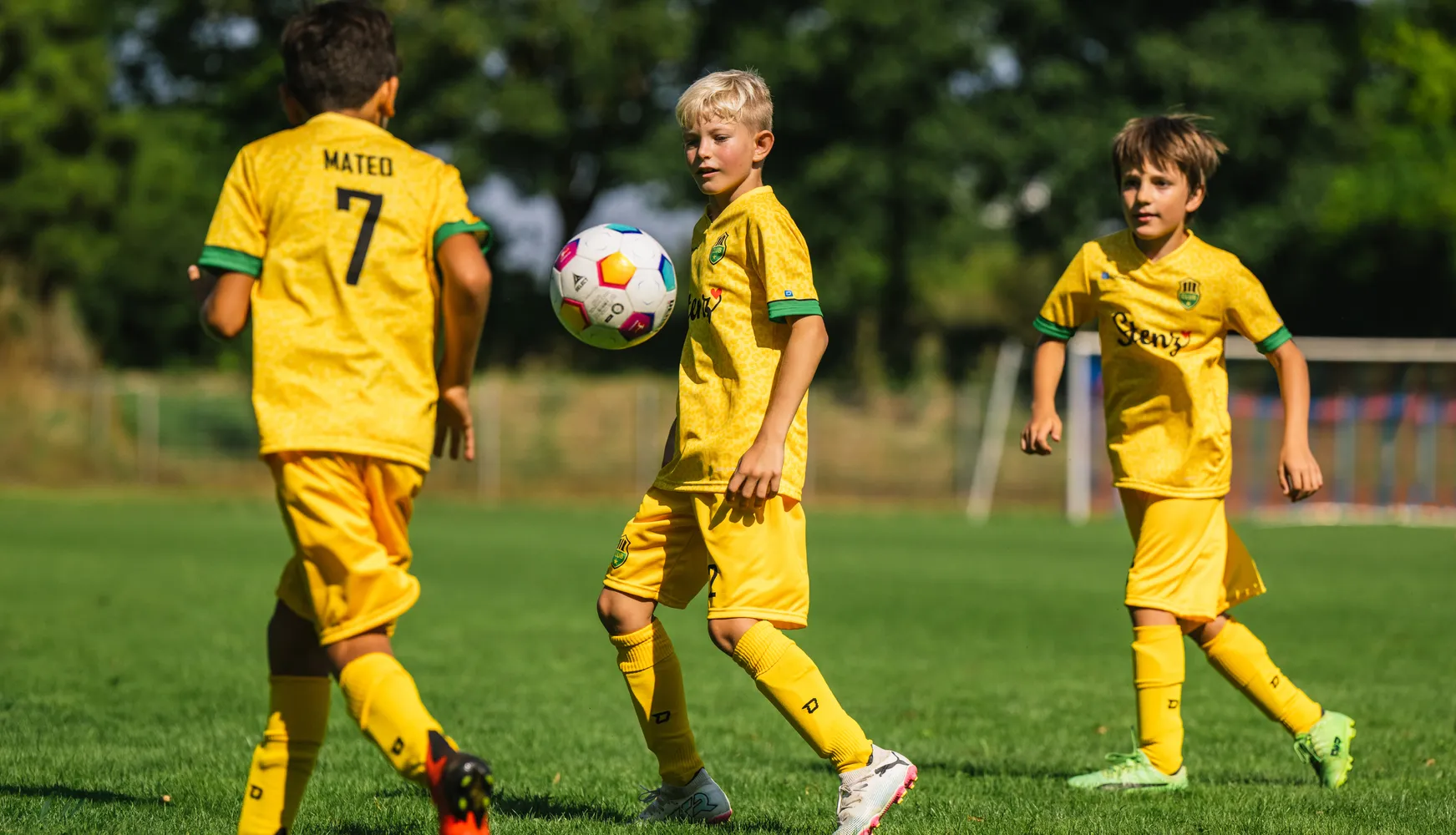 Drie kinderen in gele voetbalshirts spelen op een grasveld met een kleurrijke voetbal.