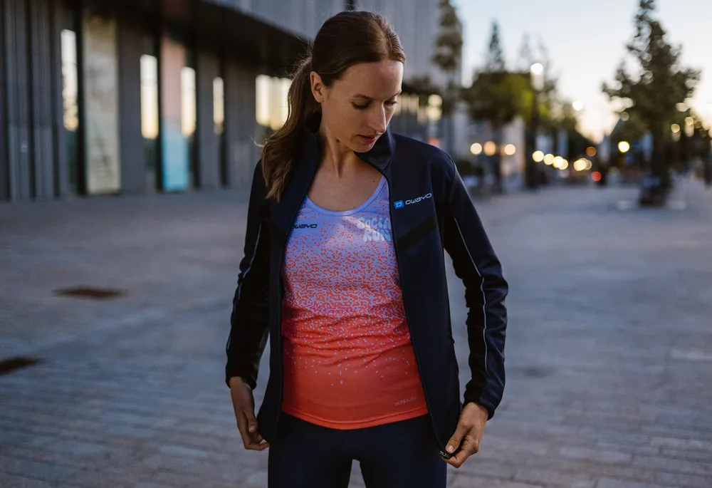 Een sportieve vrouw draagt een donkerblauwe jas over een mouwloos shirt met een blauw-oranje gradient terwijl ze in een stedelijke omgeving staat.