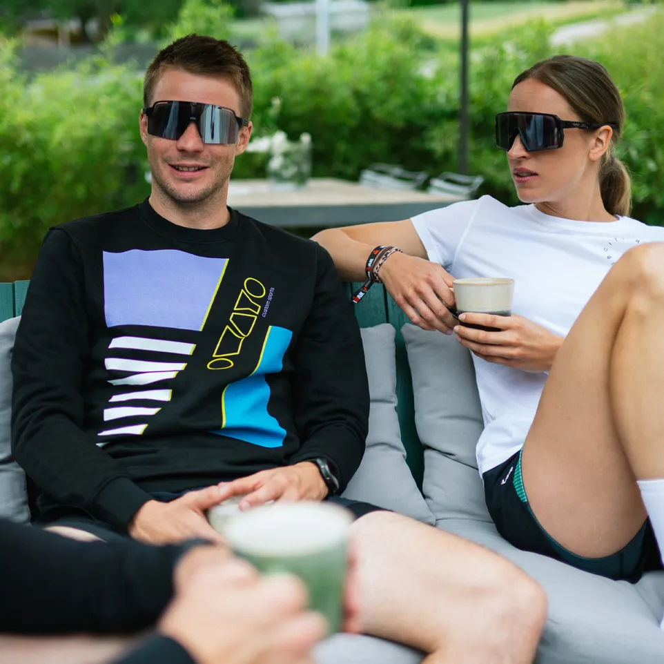 Twee mensen zitten op een buitenbank, de man draagt een zwarte sweatshirt met een kleurrijk ontwerp en de vrouw een wit t-shirt, beiden houden drankjes vast.