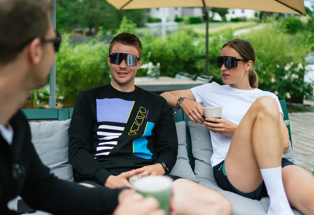 Drie mensen op een bank, twee in casual sportkleding en zonnebrillen, terwijl ze ontspannen en kletsen met drankjes in hun handen.