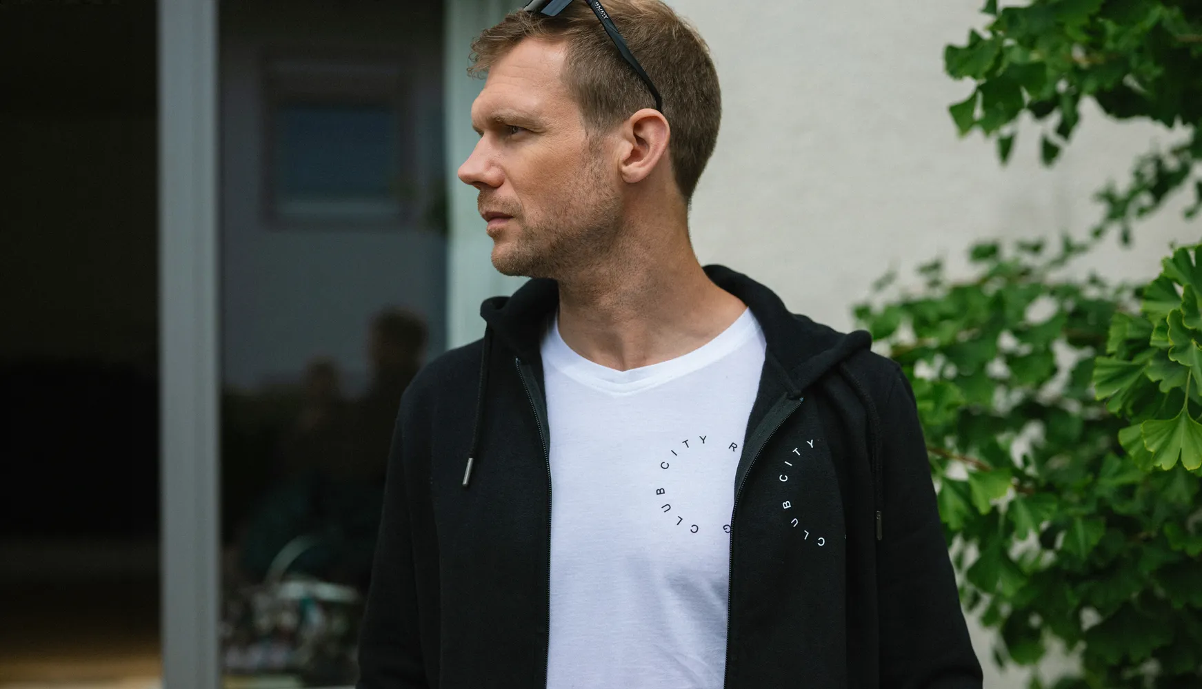 Een man in een zwart hoodie over een wit T-shirt met een cirkelvormige print staat in een buitenomgeving.