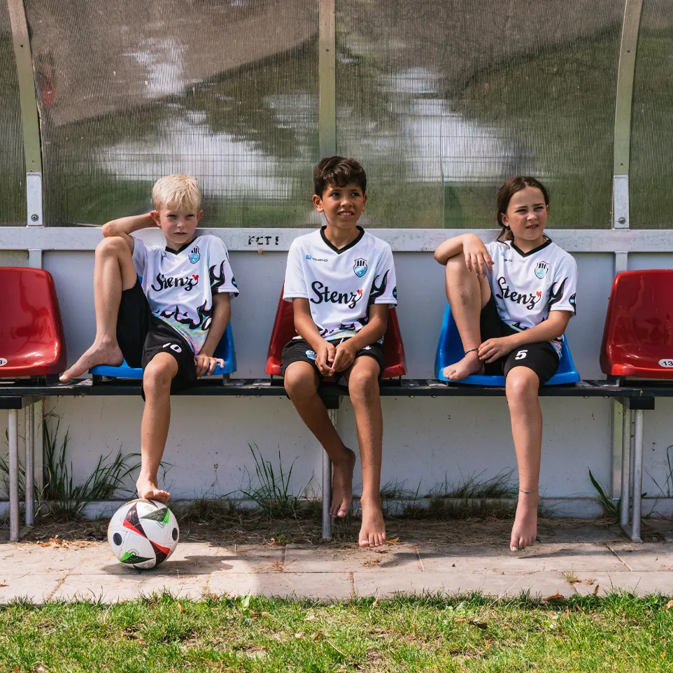 Drie kinderen in voetbalshirts en shorts zitten op een bank langs het veld, met een voetbal voor hen.