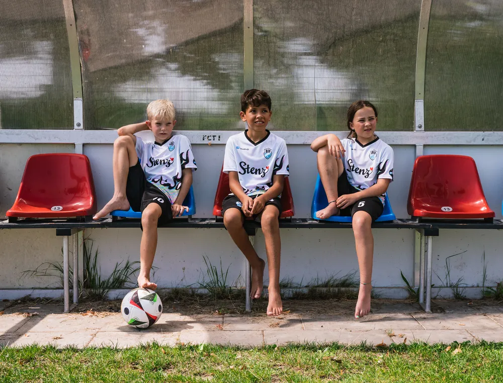 Drie kinderen zitten op een bank aan de rand van een voetbalveld, gekleed in op maat gemaakte voetbalshirts met een voetbal voor zich.