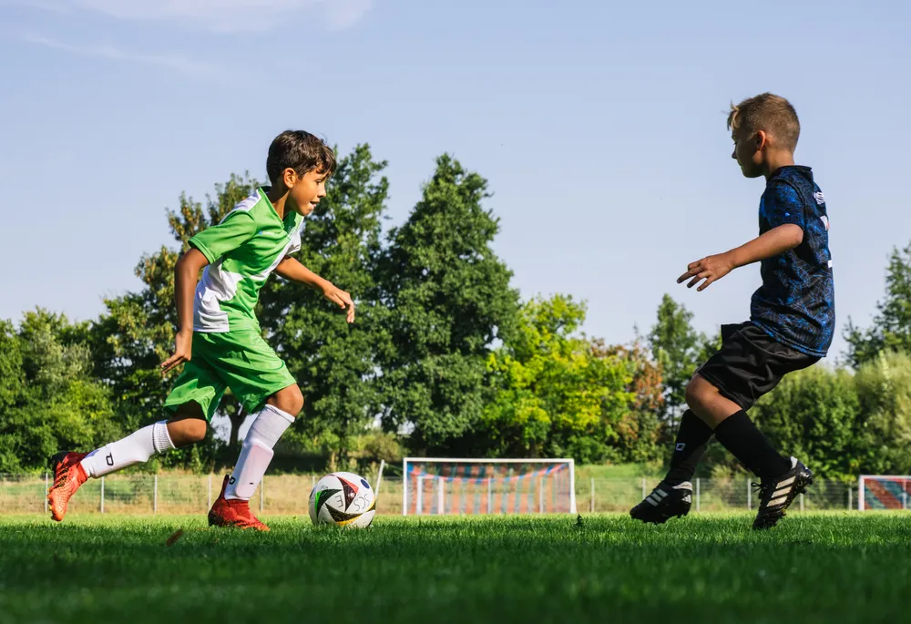 Twee kinderen spelen voetbal op een groen veld, de een in een groen shirt met witte accenten en de ander in een blauw shirt, terwijl ze achter een bal aan rennen.