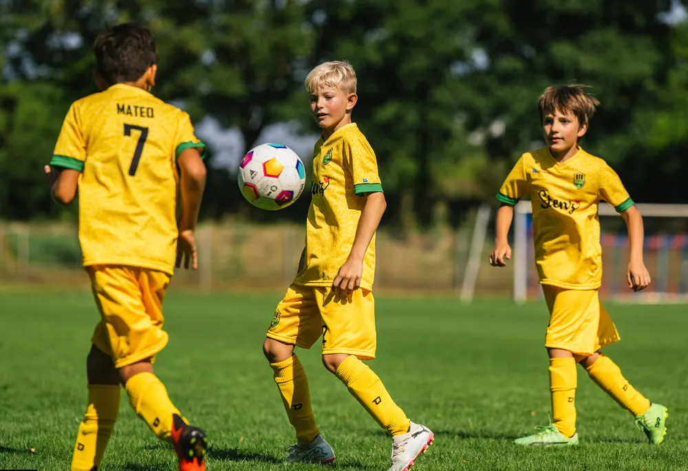 Drie kinderen in heldergele voetbalshirts spelen op een grasveld.