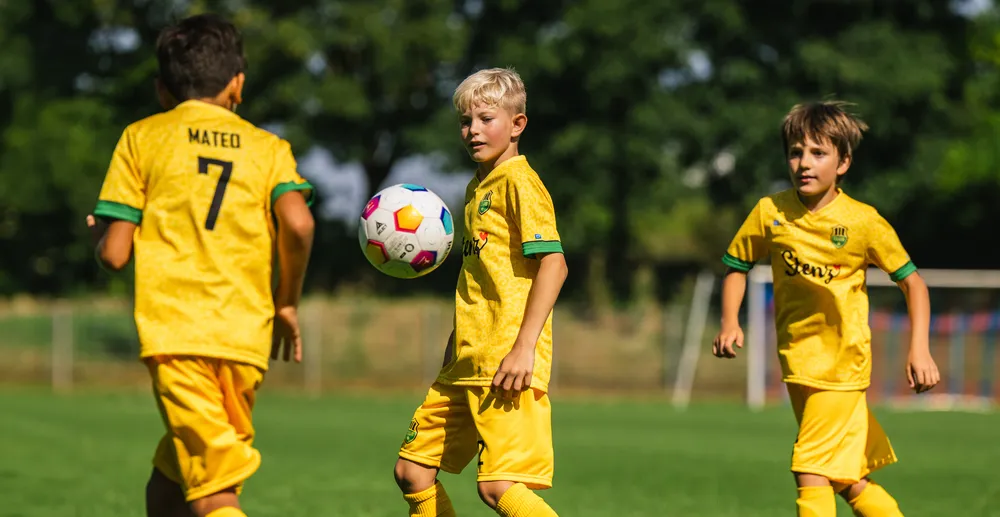 Drie kinderen in gele voetbalshirts spelen op een groen veld, terwijl een van hen een kleurrijke voetbal controleert.