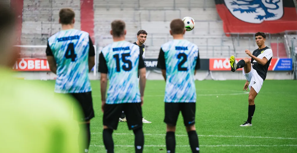 Een voetballer maakt een schot terwijl andere spelers in kleurrijke shirts toekijken.