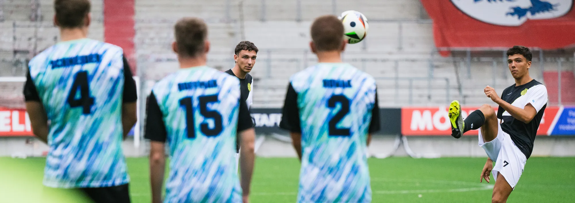 Een groep voetballers in kleurrijke shirts staat op het veld terwijl een van hen een bal met de voet optilt.