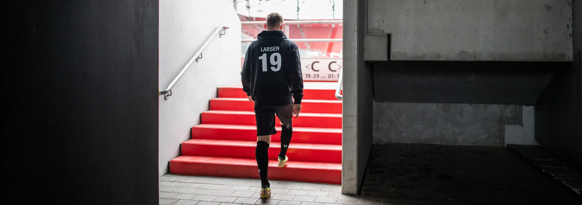 Een speler in een zwarte hoodie met het nummer 19 loopt de rode trappen uit een stadion af.