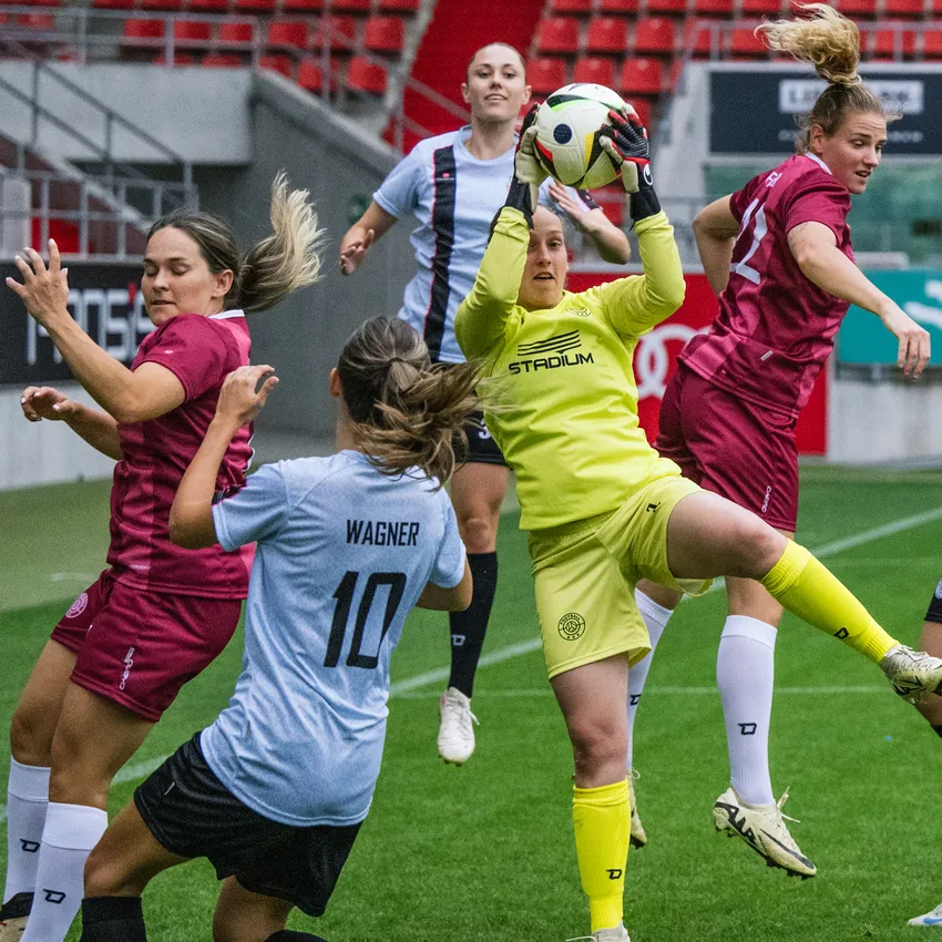 Een doelvrouw in gele sportkleding vangt de bal terwijl spelers in verschillende shirts op het voetbalveld bezig zijn.