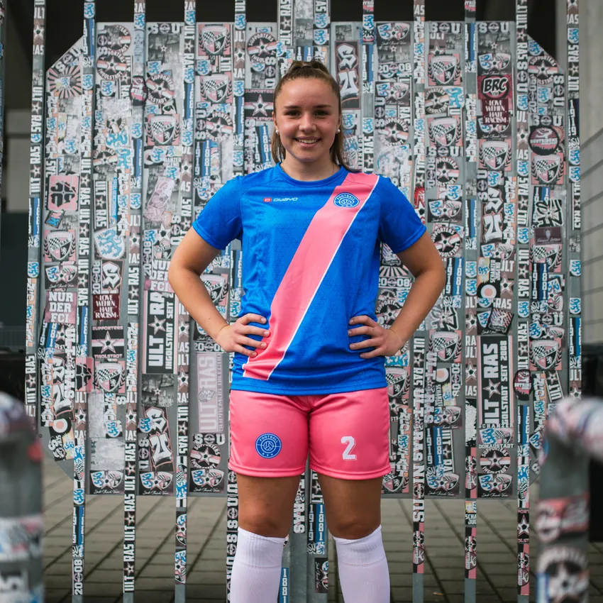 Een meisje glimlacht voor een muur vol stickers, terwijl ze een blauw voetbalshirt met een roze streep en een roze short draagt.