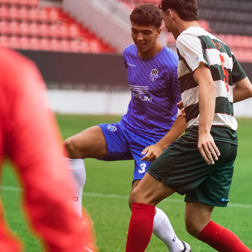 Twee voetballers in actie op het veld, de een in een blauw shirt en de ander in een groen-rood gestreept shirt.