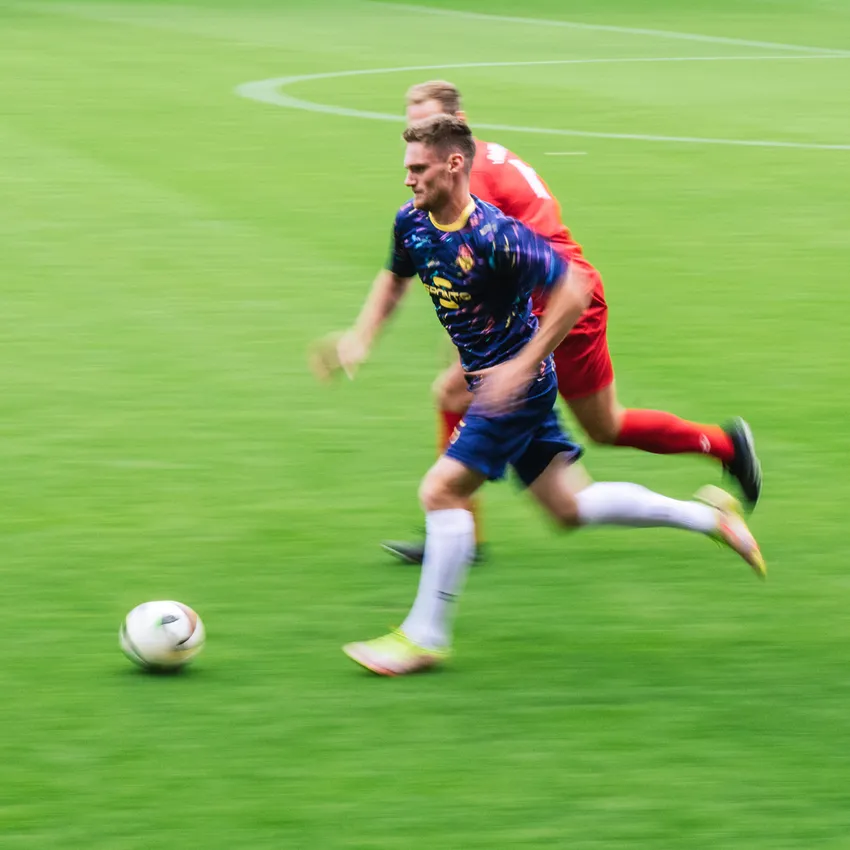 Een voetballer in een kleurrijk shirt sprint over het groene veld terwijl een andere speler hem achtervolgt.