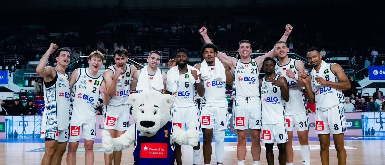 Basketbalspelers in witte shirts met het logo van BLG Logistics vieren op het veld, omringd door een publiek, met een ijsbeer-mascotte op de voorgrond.